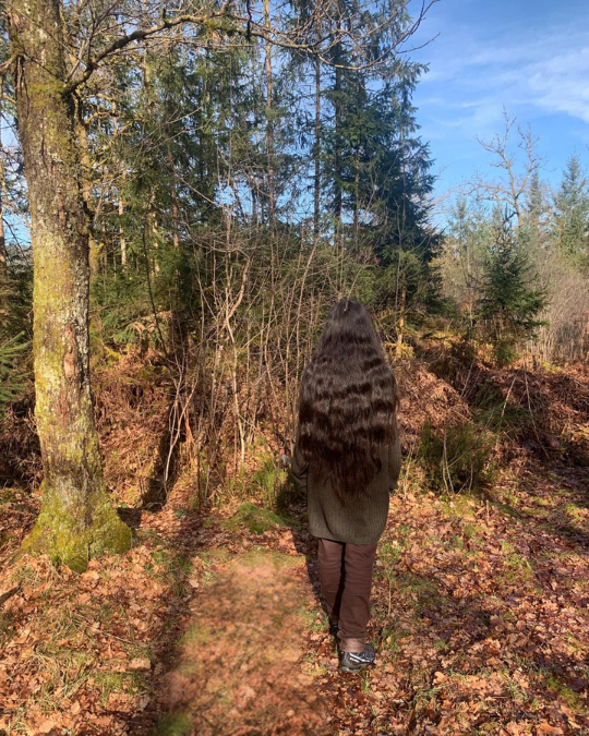Femme de dos marchant sur un sentier forestier baigné de lumière naturelle, entourée d’arbres et de végétation.
